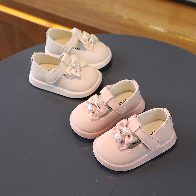 Two pairs of toddler shoes on a dark surface, one white and one pink. Each has a bow and pearl decoration on the strap.