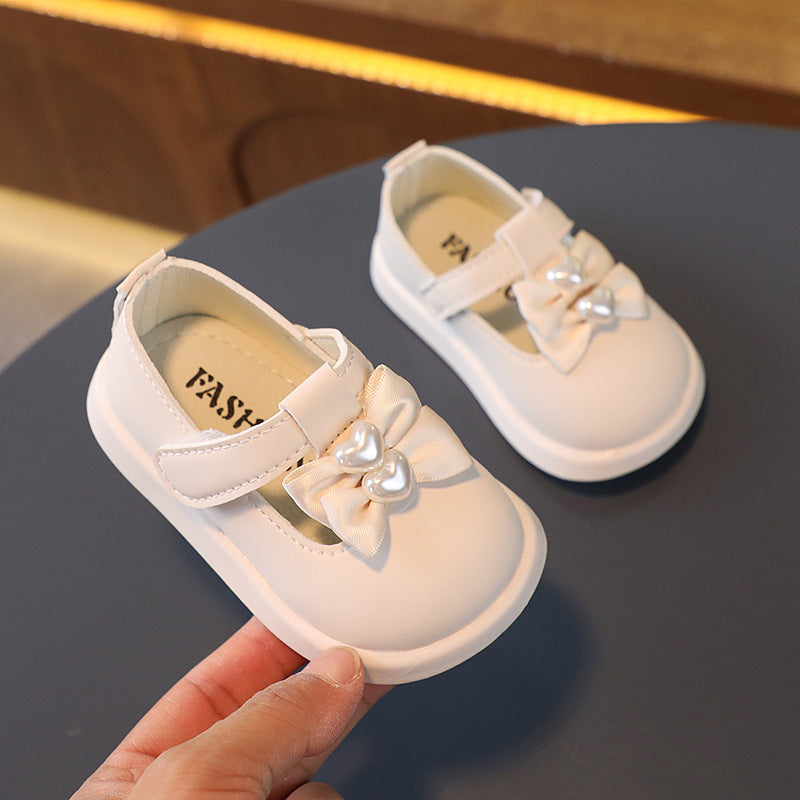 A pair of white infant shoes with Velcro straps and decorative bows adorned with heart-shaped pearls, displayed on a dark surface. One shoe is held up for a closer view.