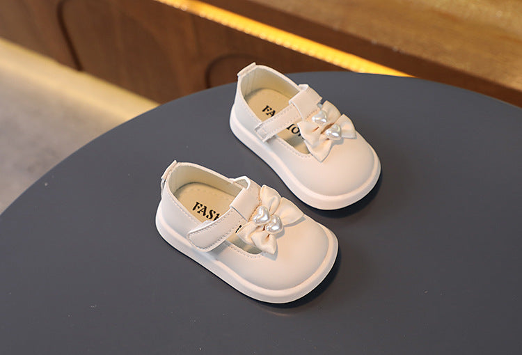 Pair of white baby shoes with decorative bows and pearls on a round gray table. The shoes have Velcro straps, and the background includes a wooden surface with warm lighting.