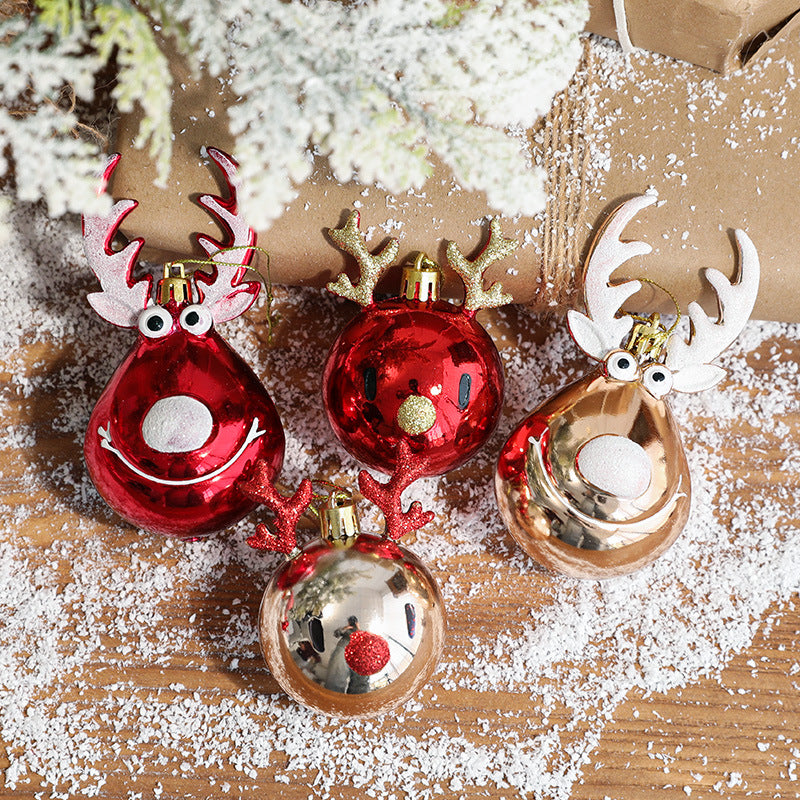Festive reindeer-shaped ornaments in red and gold rest on a wooden surface dusted with snow-like sprinkles, next to wrapped gifts and snow-covered greenery.