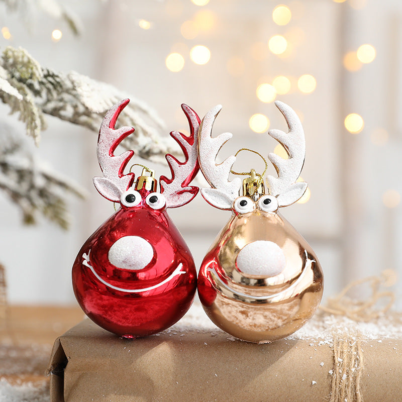 Two festive reindeer ornaments with large eyes and antlers, one red and one gold, sit on a snowy gift box. Soft lights and snow-dusted branches create a cozy holiday scene.