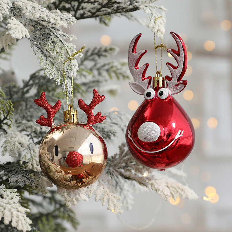 Two festive reindeer-themed ornaments hang on a snow-dusted tree branch. One is gold with red antlers and nose; the other is red with silver antlers and a smile.
