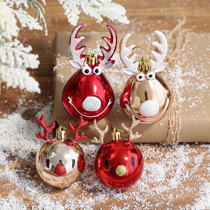 Shiny reindeer ornaments in red and gold with playful eyes and antlers, placed on a snow-dusted wooden surface with a wrapped gift and snowy pine branches in the background.