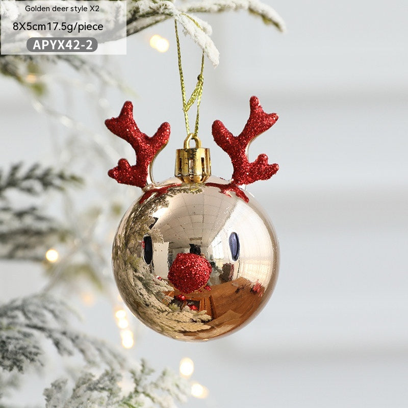 Golden Christmas ornament shaped like a reindeer with red glitter antlers and nose, hanging on a snow-dusted tree branch. Reflections visible on its shiny surface.