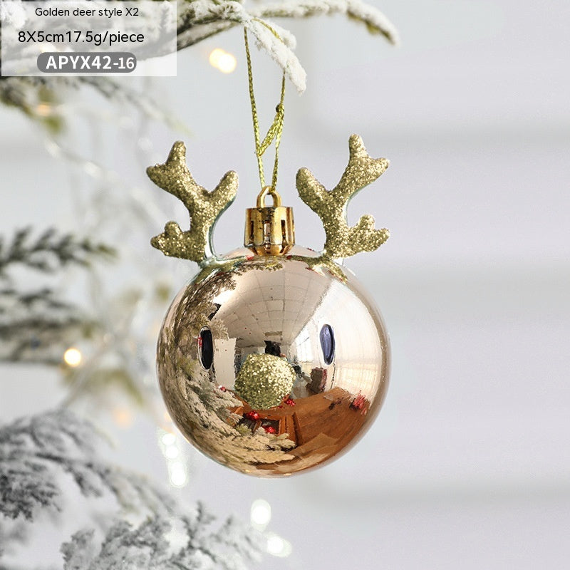 A shiny gold Christmas ornament shaped like a reindeer face, complete with glittery antlers, hangs from a snow-dusted tree branch. Reflections are visible on its surface.