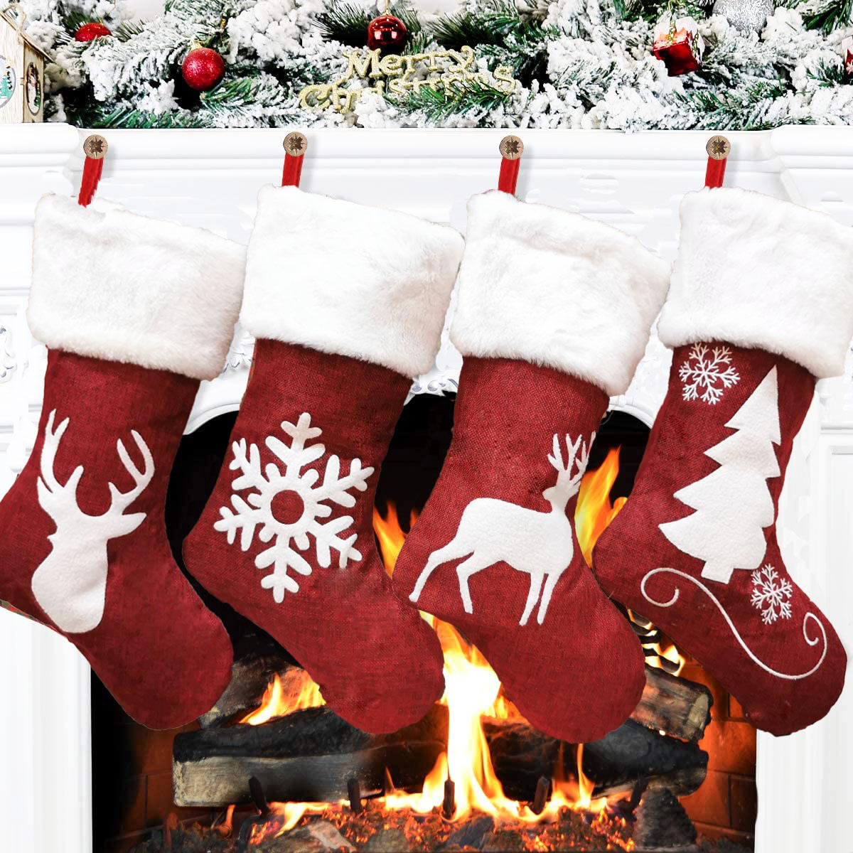 Four red Christmas stockings with white fur trim hang on a mantel. Each stocking has a unique festive design: reindeer, snowflake, stag, and tree. A glowing fire burns below.