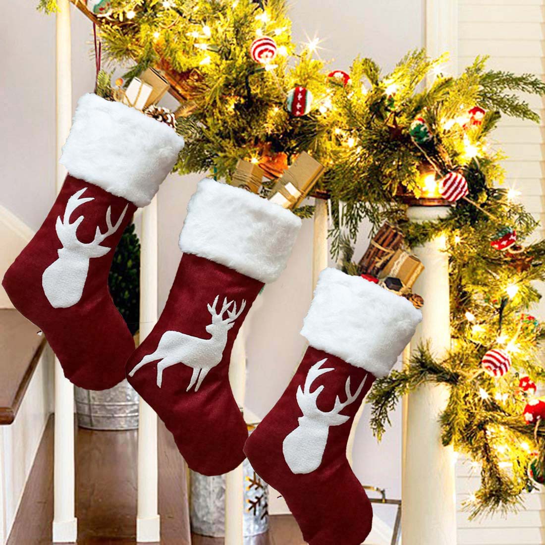 Three red stockings with white cuffs and deer patterns hang on a staircase railing adorned with green garland, string lights, and colorful ornaments.