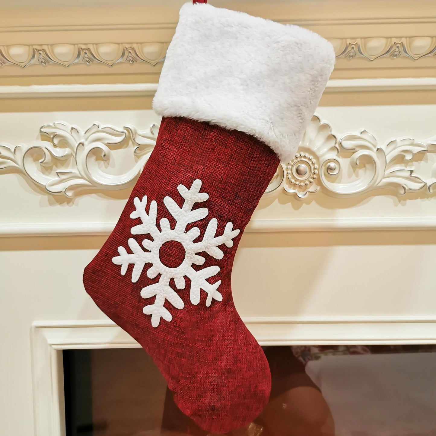 Red Christmas stocking with a white snowflake design, hanging on an ornate fireplace. The stocking has a fluffy white cuff and intricate white floral detailing on the mantel.