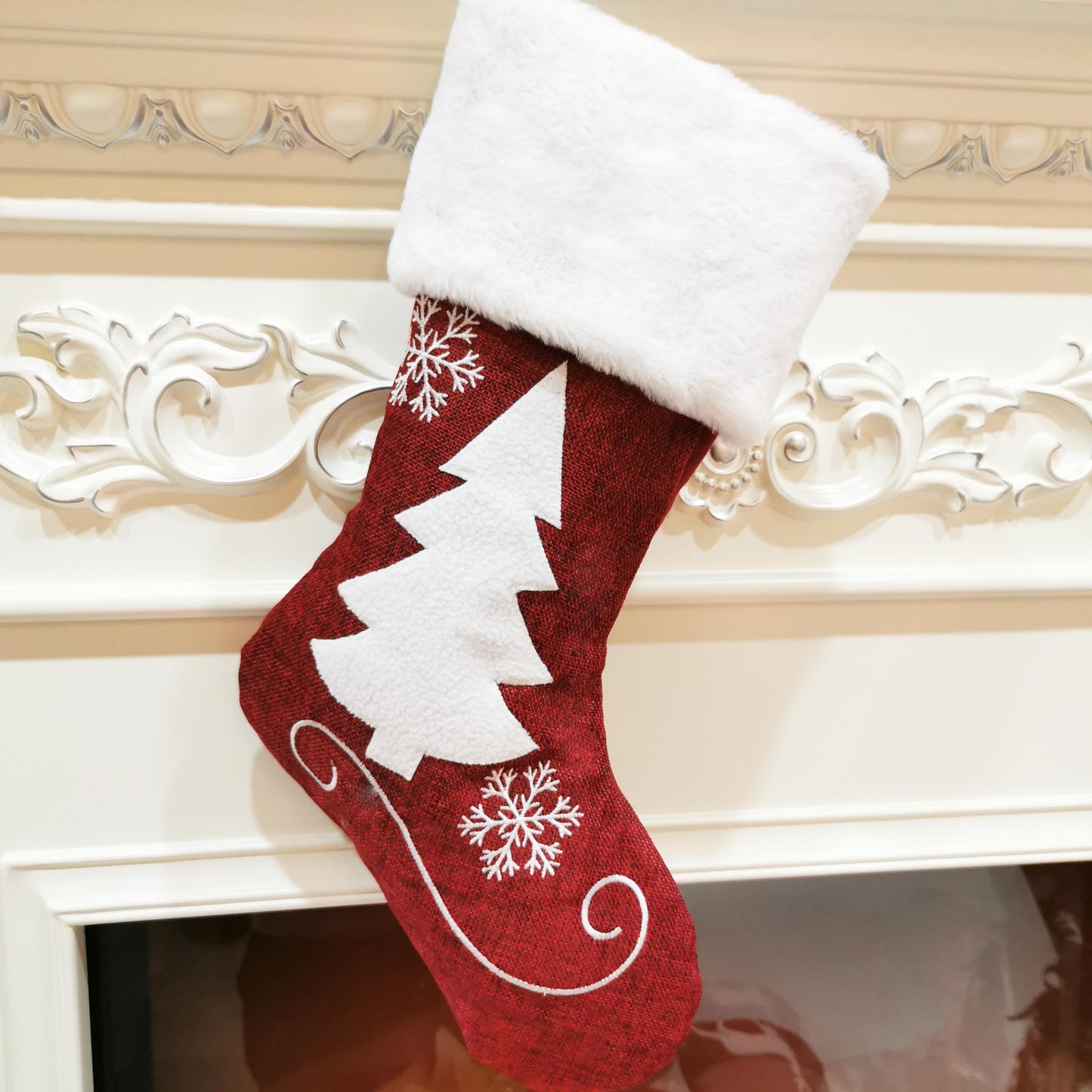 A red Christmas stocking with white fur trim hangs on a decorative mantle. It features a white tree and snowflakes design.
