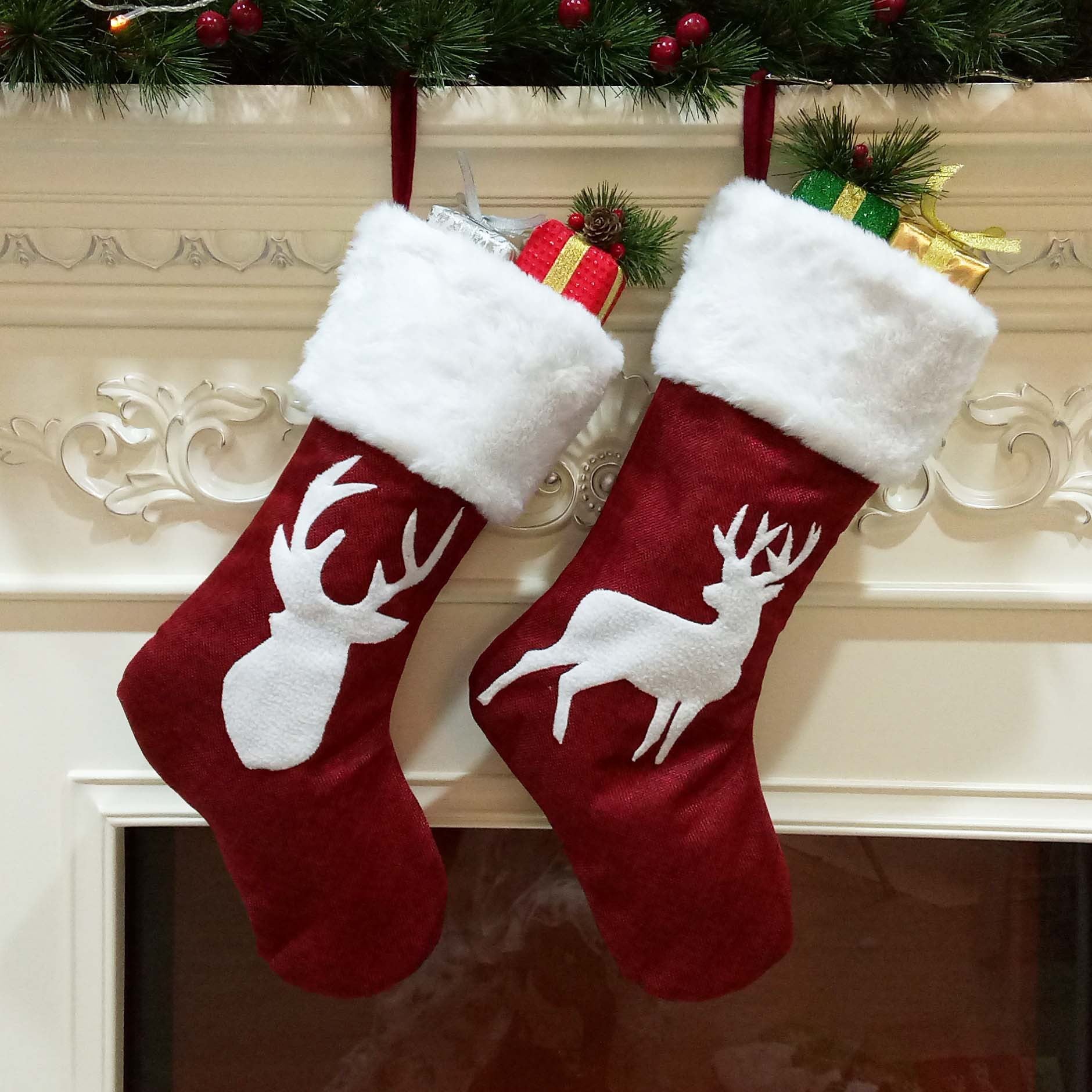 Two red Christmas stockings with white deer motifs hang on a decorative mantel. Each is filled with wrapped gifts, adorned with evergreen sprigs and festive accents.