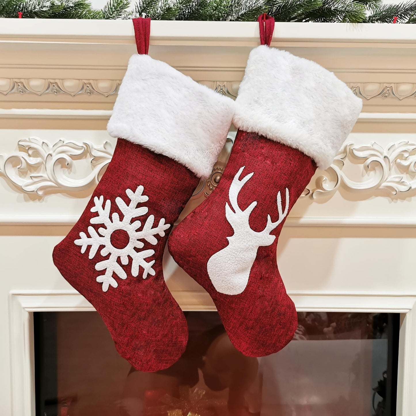 Two red Christmas stockings with white cuffs hang on a decorated mantel. One features a snowflake design, and the other shows a reindeer silhouette.