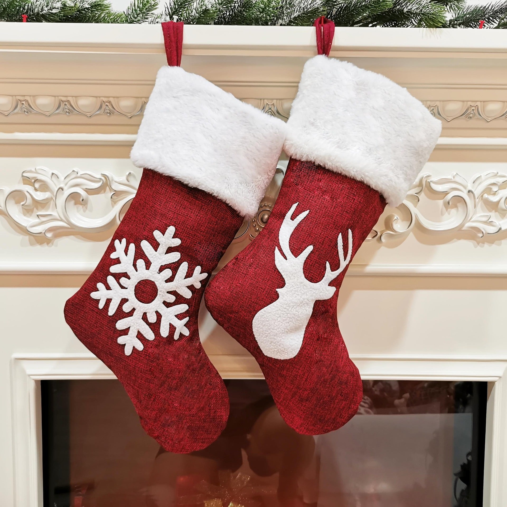 Two red Christmas stockings with white cuffs hang on a decorated mantel. One features a snowflake design, and the other shows a reindeer silhouette.