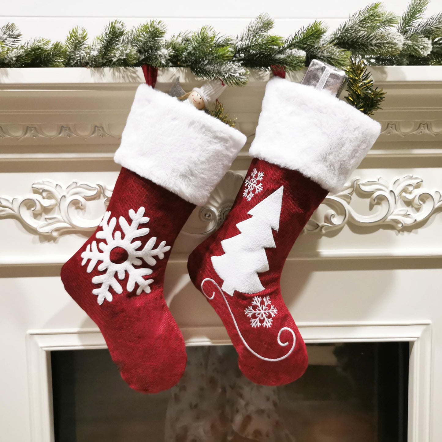 Two red Christmas stockings with white trim hang on a decorated mantle. One features a snowflake design, the other a tree. Gifts and greenery peek out from the tops.