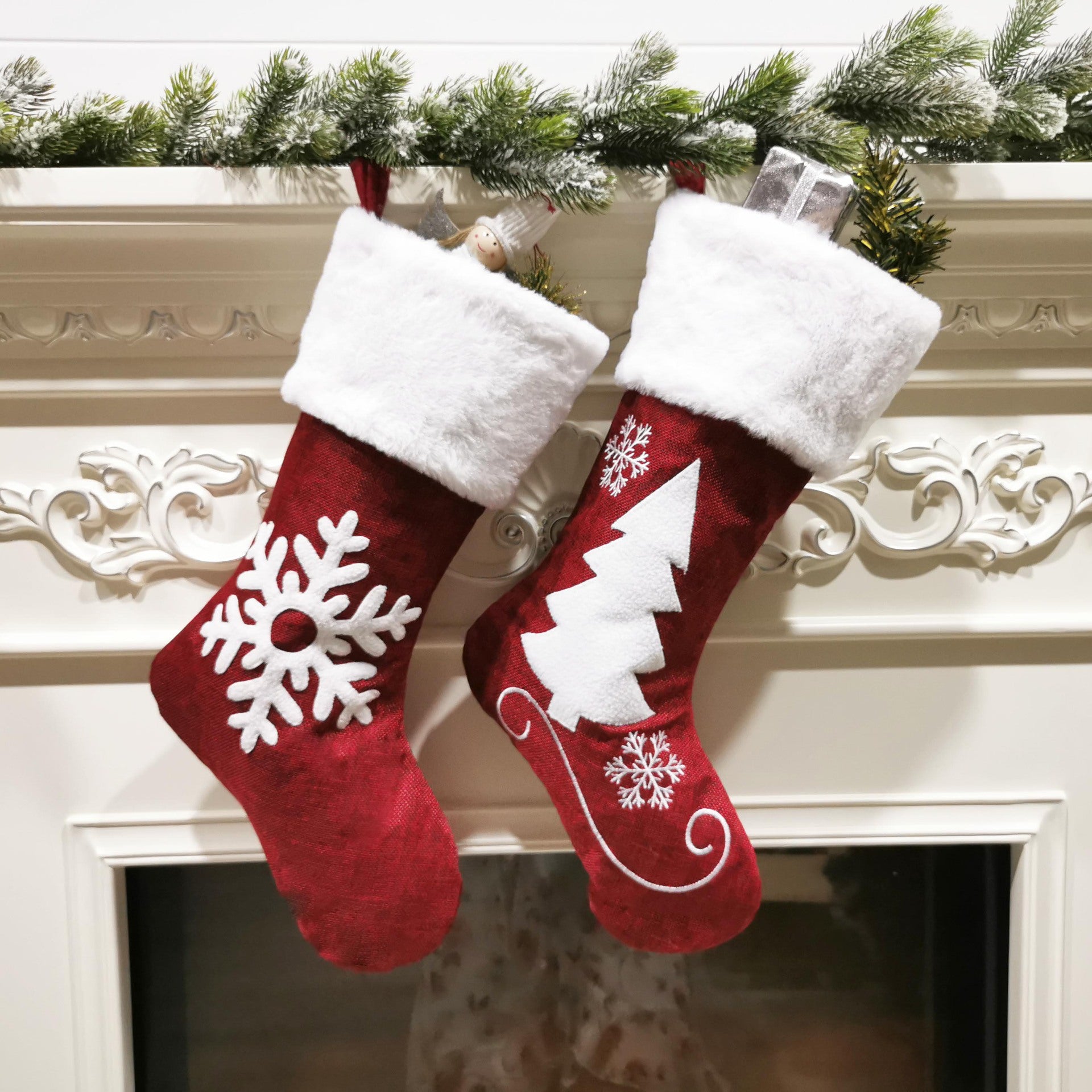 Two red Christmas stockings with white trim hang on a decorated mantle. One features a snowflake design, the other a tree. Gifts and greenery peek out from the tops.