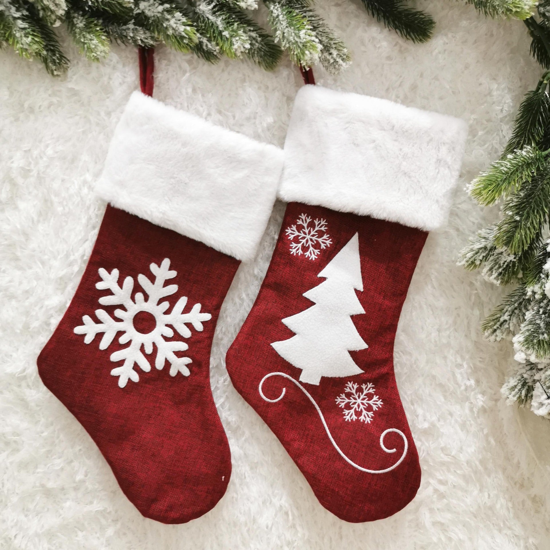 Two red Christmas stockings with white cuffs hang on a snowy background. One has a snowflake, and the other a tree and snowflakes. Pine branches frame the top corners.