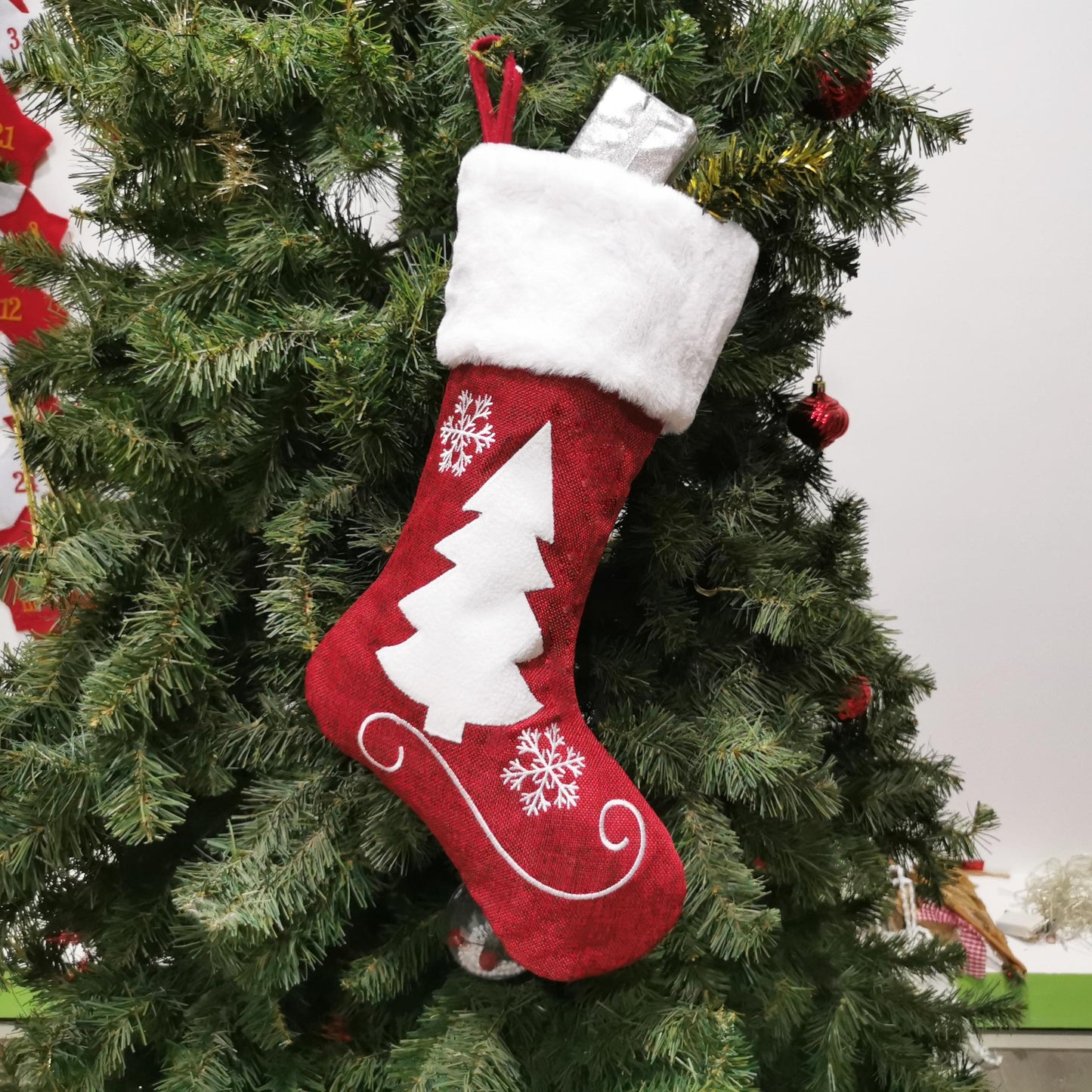 A red Christmas stocking with a white tree and snowflakes hangs on a green tree, filled with a silver wrapped gift.