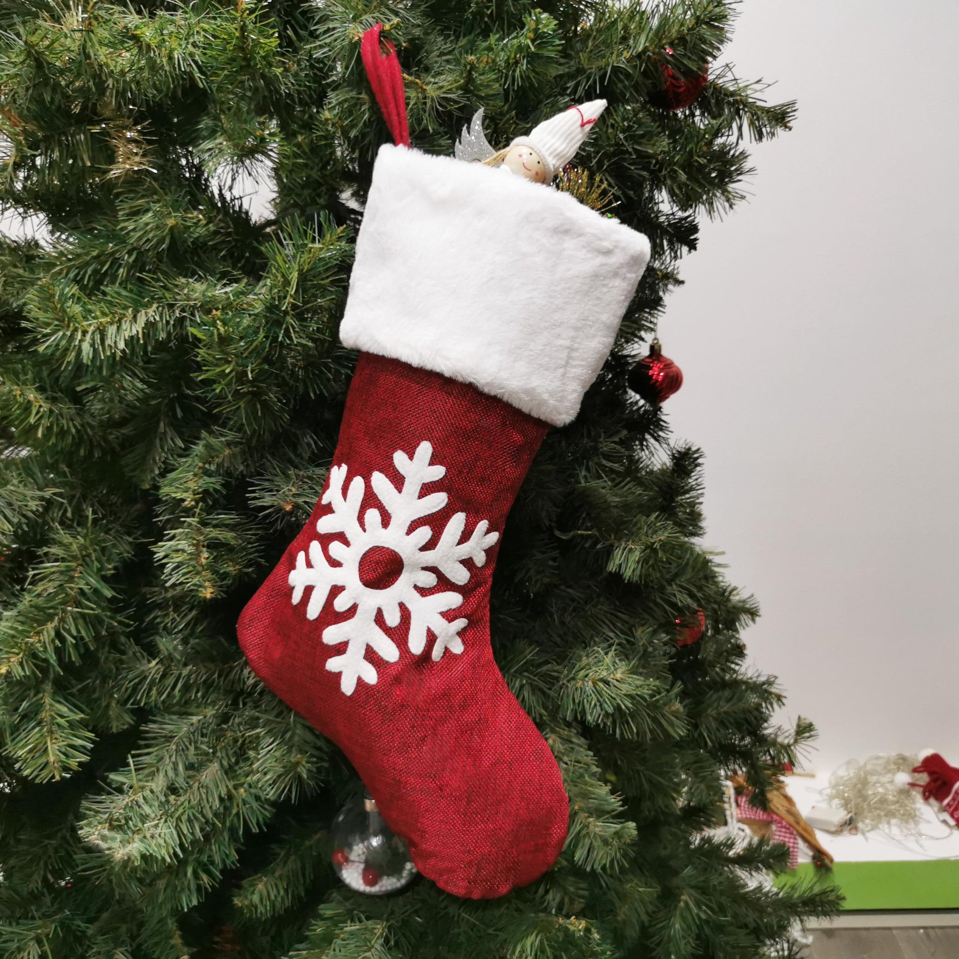 A red Christmas stocking with a white cuff and snowflake design hangs on a green tree. An angel figurine peeks out from the top, surrounded by red baubles.