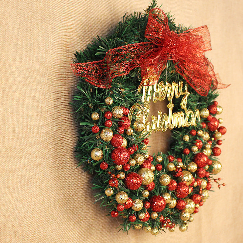 A festive Christmas wreath decorated with red and gold ornaments, a glittery "Merry Christmas" sign, and a red mesh bow, hangs on a beige wall.