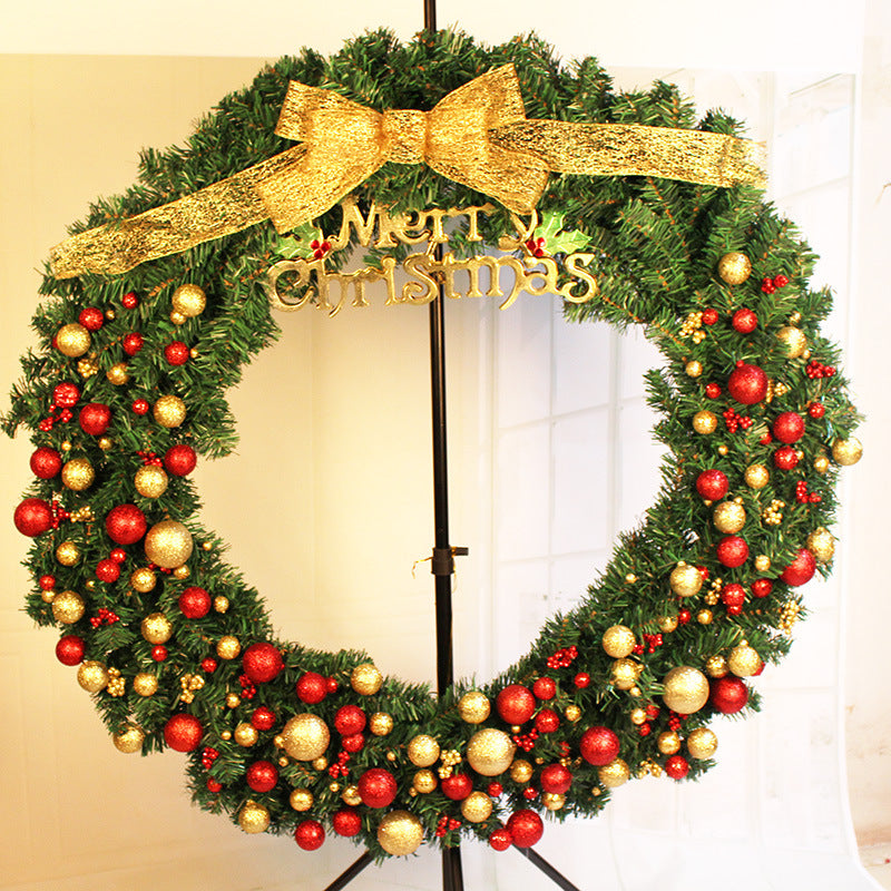 A large Christmas wreath decorated with red and gold baubles, adorned with a big gold bow and a "Merry Christmas" sign. It is displayed on a black stand indoors.