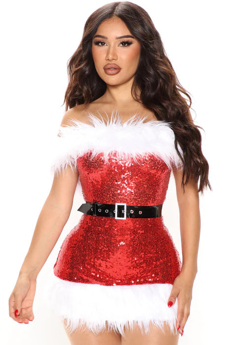 Woman in a sparkly red off-shoulder Santa-inspired dress with white fur trim and black belt. Long wavy brown hair, bold makeup, standing against a white background.
