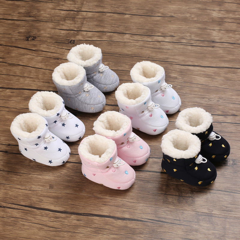 Cozy baby booties with soft interiors are arranged in pairs on a wooden floor. Designs include gray with stars, white with blue stars, pink with blue dots, and black with gold stars.