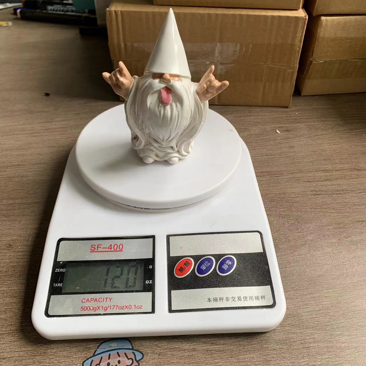 A ceramic gnome with a pointed hat and beard, making a playful gesture, sits on a digital scale reading 120 grams, surrounded by cardboard boxes on a wooden surface.
