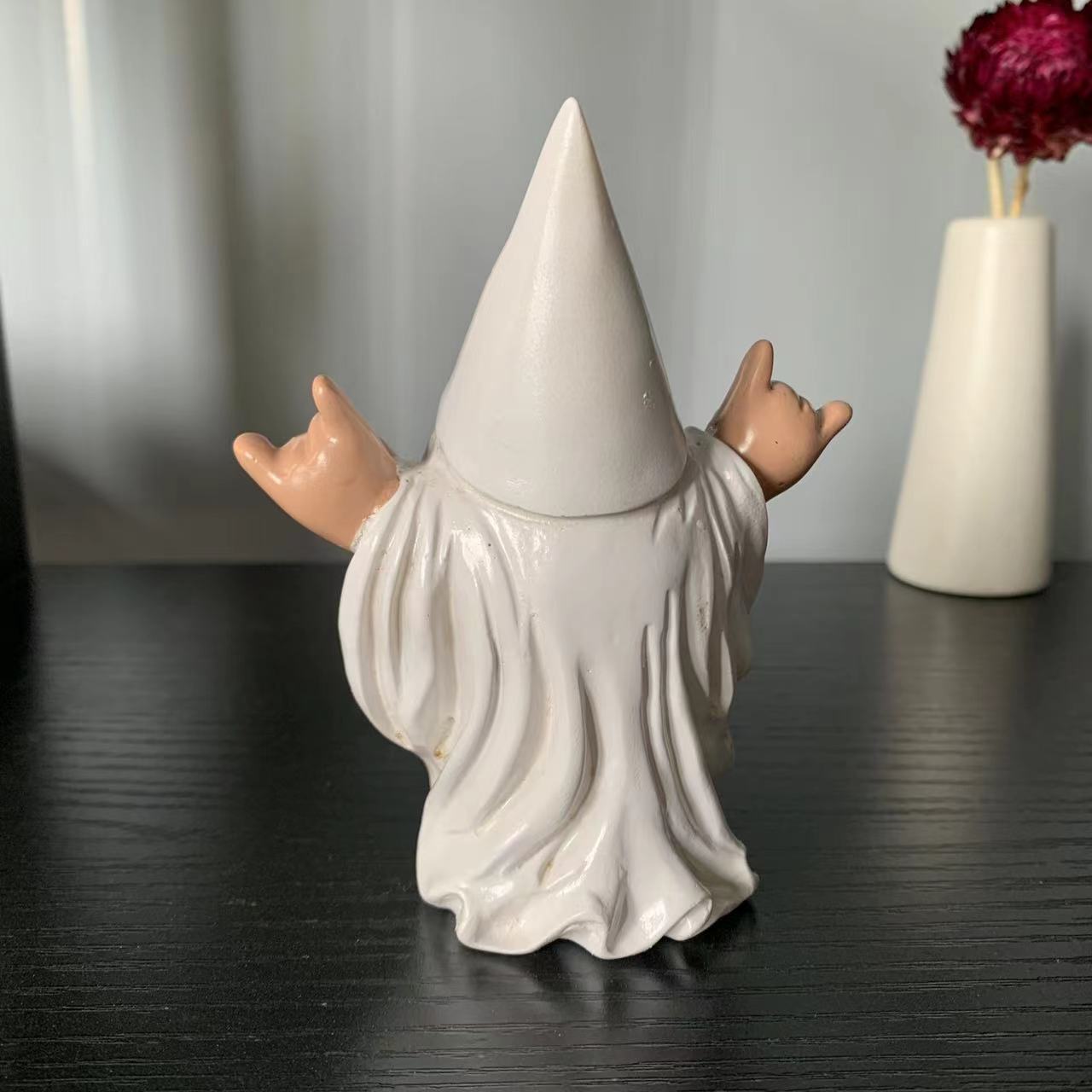A small figurine with a tall pointed hat and flowing robe raises its hands in a rock and roll gesture. It's placed on a dark surface with a decorative vase in the background.