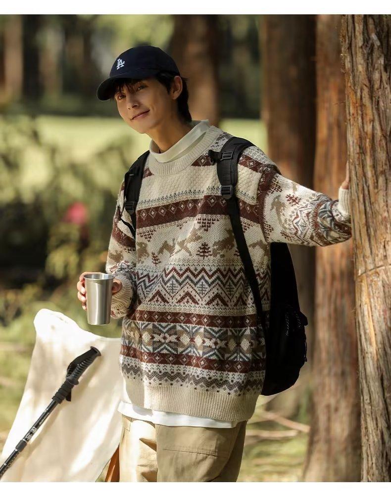 A person in a patterned sweater and cap stands outdoors, holding a metal cup and leaning against a tree with a backpack, in a forest setting.