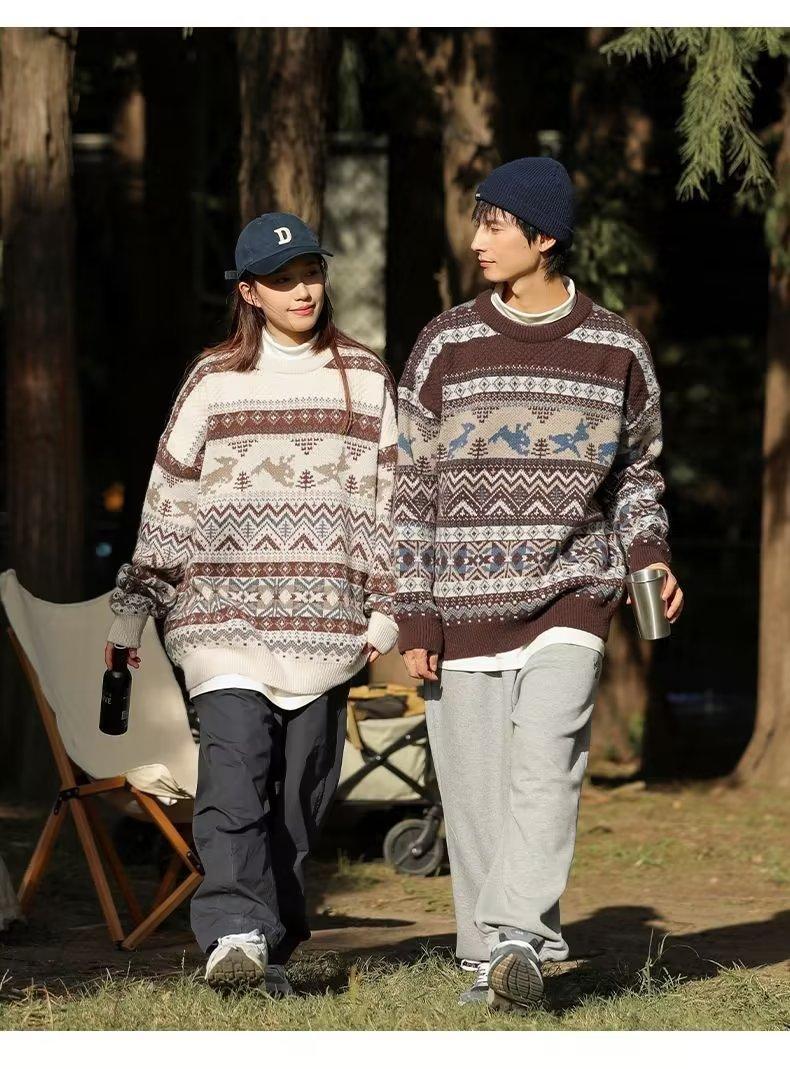 A couple walks in a wooded area, both wearing patterned sweaters and hats. The woman holds a bottle while the man holds a cup. They appear to be enjoying a casual stroll.