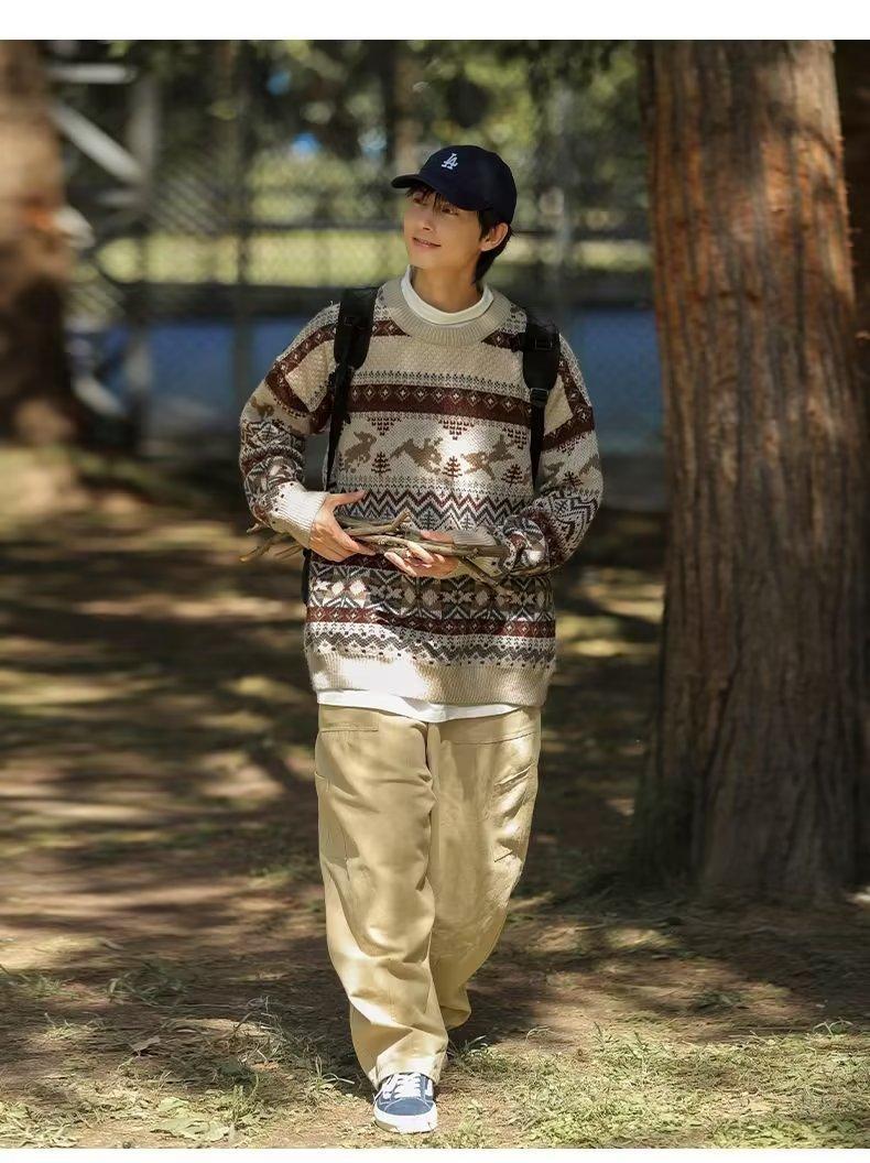 A person wearing a patterned sweater, beige pants, and a black cap carries sticks while walking in a sunlit, wooded area. A backpack is slung over their shoulders.