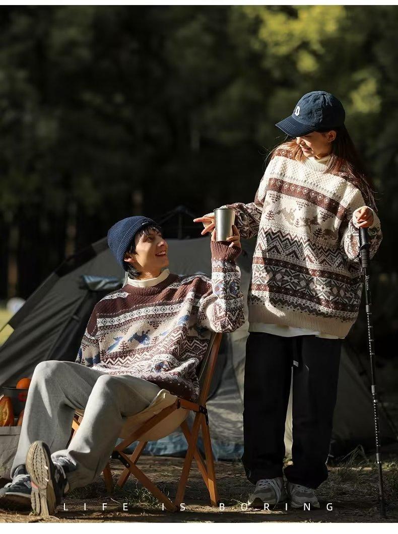 Two people in cozy patterned sweaters and hats enjoy a camping moment. One sits on a chair holding a cup, while the other stands smiling with a walking stick. A tent is in the background.