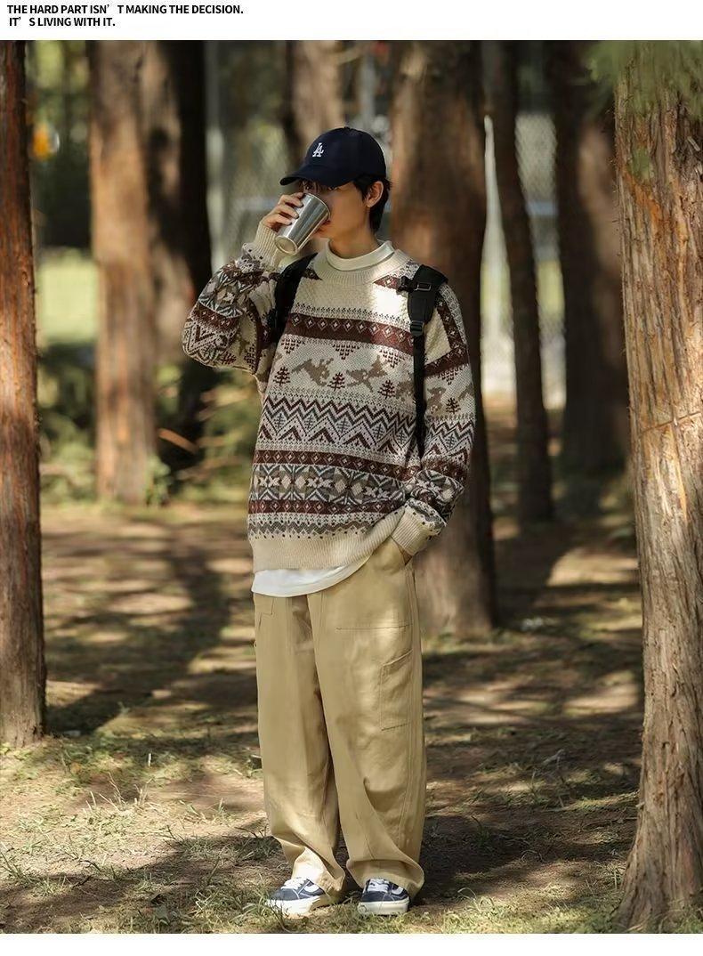 Person in a patterned sweater, khaki pants, and baseball cap drinks from a cup while standing in a wooded area with sunlight filtering through the trees.