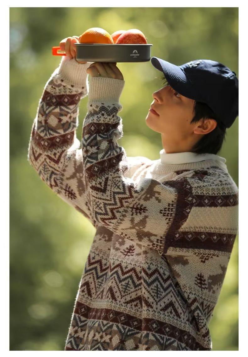 A person in a patterned sweater and cap holds a tray with oranges against a blurred green background, looking upwards contemplatively.
