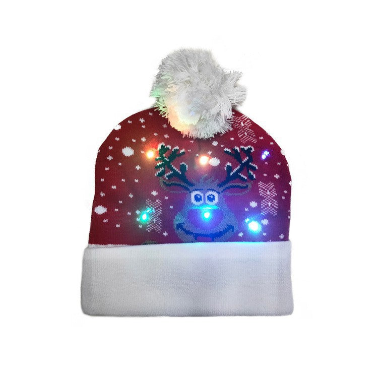 Red beanie with a reindeer pattern, adorned with colorful LED lights. It has a white fluffy pom-pom on top and a white cuff at the bottom.
