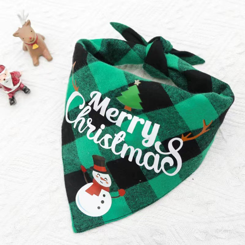 Green and black checkered dog bandana with "Merry Christmas," a snowman, and a tree design. Mini Santa and reindeer figurines on a textured white background.