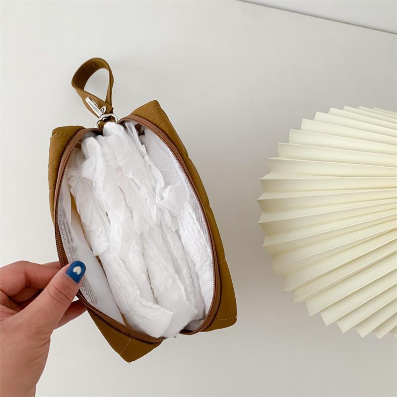 A hand with blue nail polish holds an open brown pouch filled with white crinkled items, next to a folded cream-colored paper fan on a white surface.