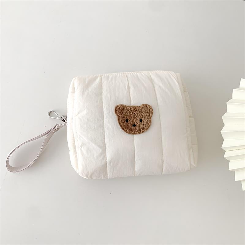 A small, quilted white pouch with a teddy bear patch in the center, featuring a wrist strap and resting on a white surface next to a folded white item on the right.