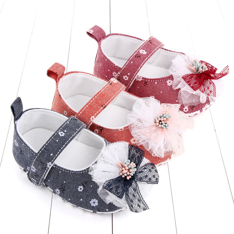 Three baby shoes with floral lace designs in pink, red, and navy blue. Each shoe has a strap and is decorated with a large tulle flower and beaded center on a white wooden surface.