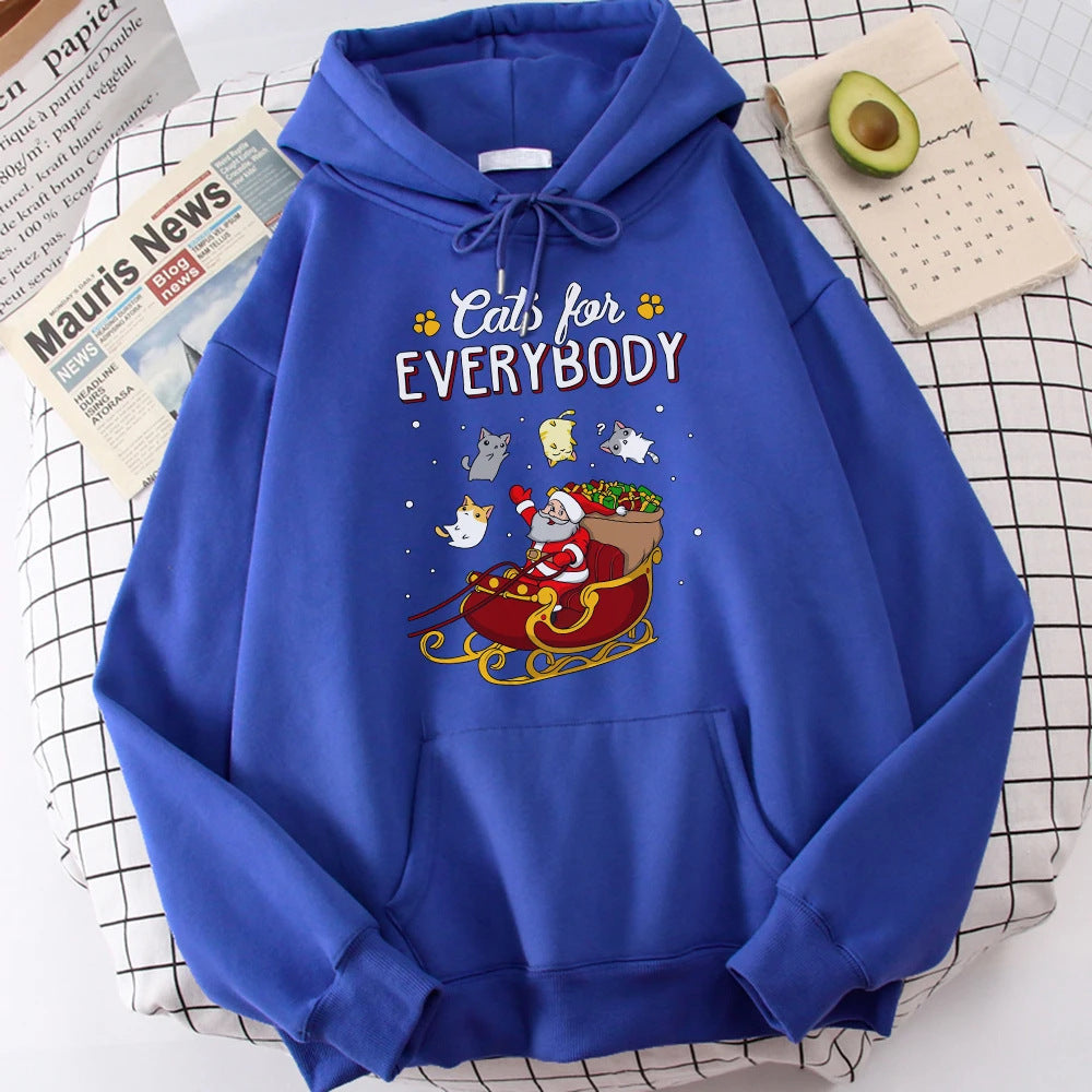 Blue hoodie with "Cats for Everybody" text and illustration of Santa in a sleigh surrounded by cats. Beside it are a newspaper, an avocado, and a small calendar.