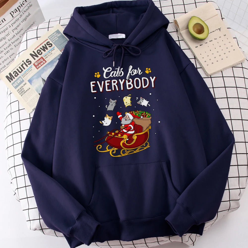 Navy hoodie on a checkered surface, featuring Santa in a sleigh surrounded by cats, with the phrase "Cats for Everybody." Nearby are a newspaper, half an avocado, and a notepad.