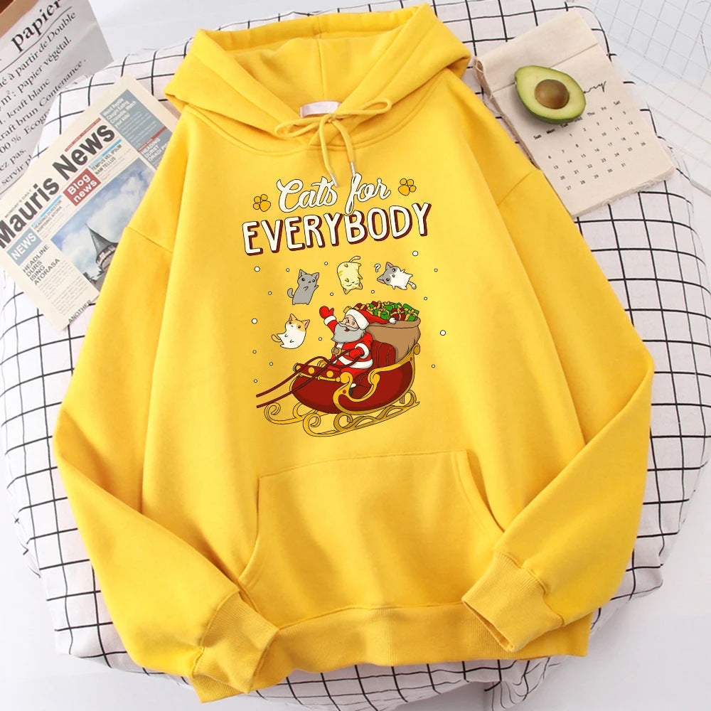 Yellow hoodie with "Cats for Everybody" text. Features Santa in a sleigh with colorful cats floating around. Background includes an avocado, newspaper, and grid-patterned cloth.