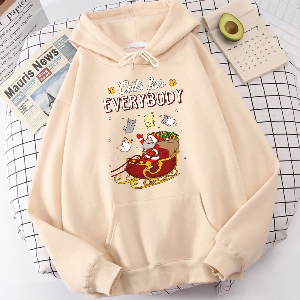 Cream hoodie with "Cats for Everybody" text featuring a cartoon Santa in a sleigh surrounded by festive cats. Background includes an avocado, newspaper, and calendar.