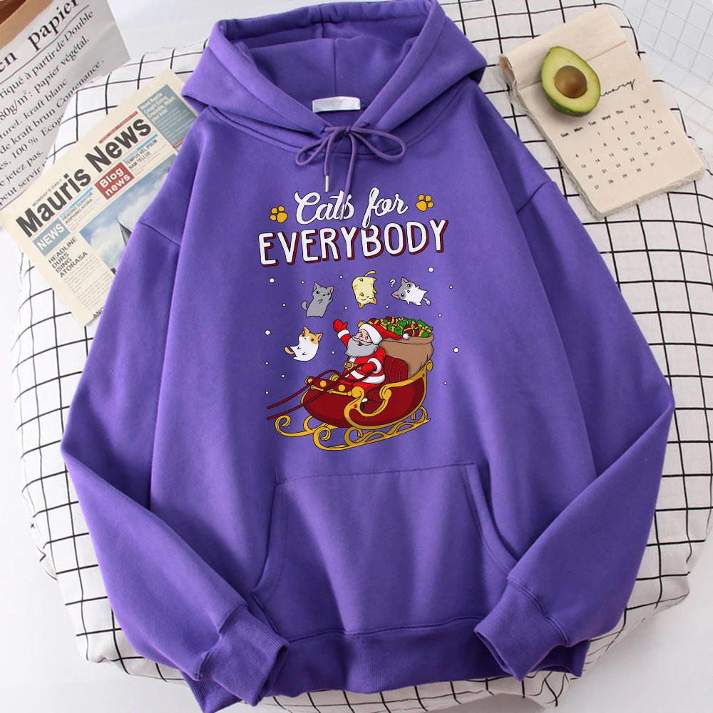 Purple hoodie with a festive design featuring Santa Claus in a sleigh surrounded by cats. Text reads "Cats for Everybody." Avocado, calendar, and newspaper in the background.