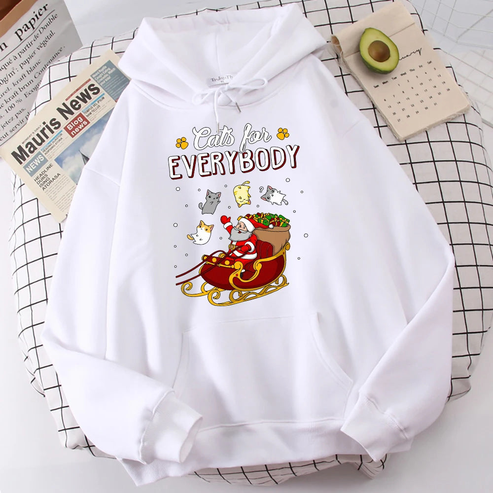White hoodie with "Cats for Everybody" text and a design of a Santa in a sleigh, surrounded by playful cats. Nearby, there's a newspaper, a calendar, and a sliced avocado.