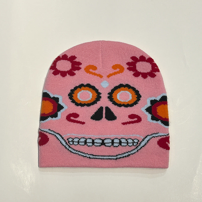 A pink beanie featuring a colorful Día de los Muertos sugar skull design with floral patterns, vibrant orange and red accents, and a stylized smiling mouth on a plain background.