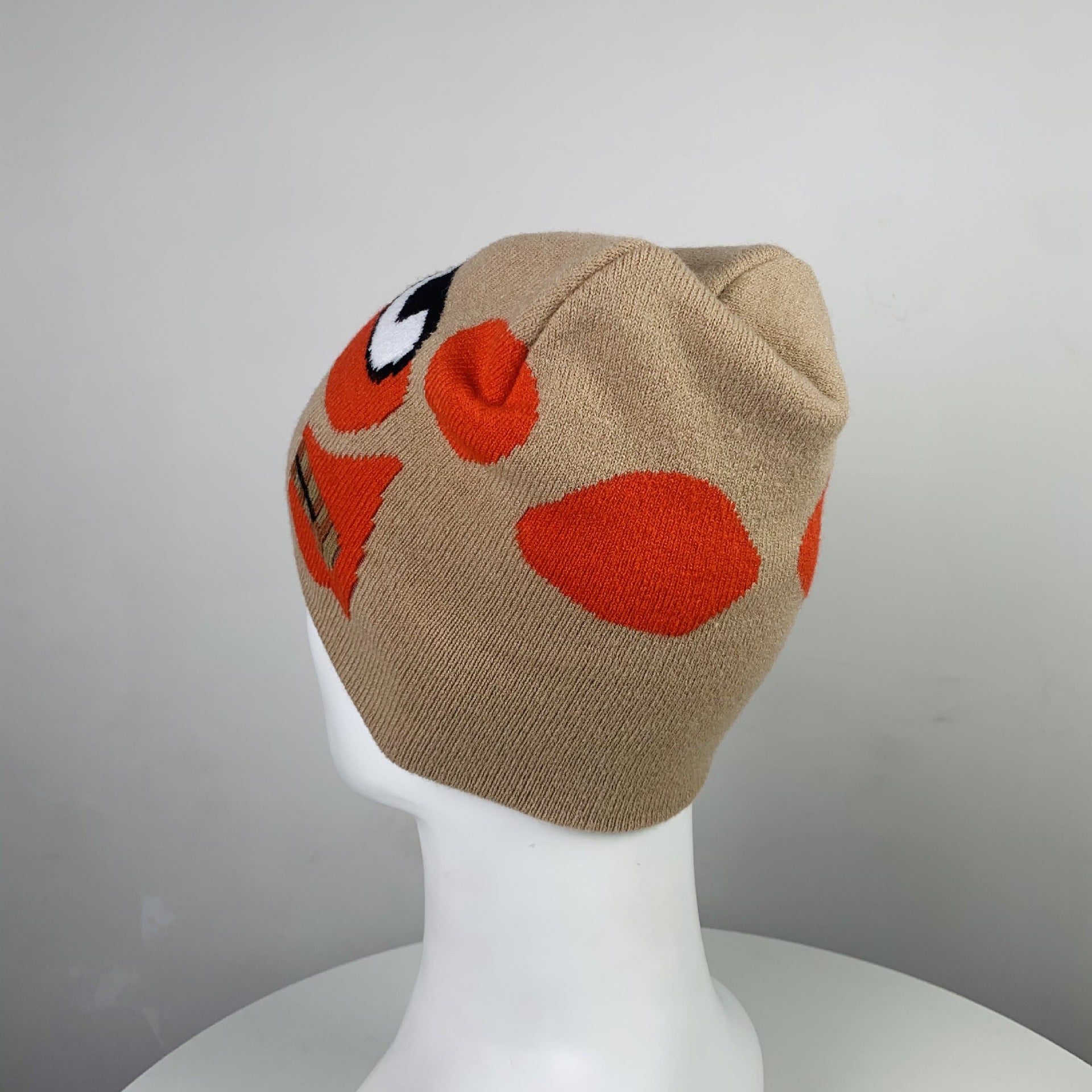 Tan knit beanie with bright orange and black cartoon face design, displayed on a white mannequin head against a neutral background.