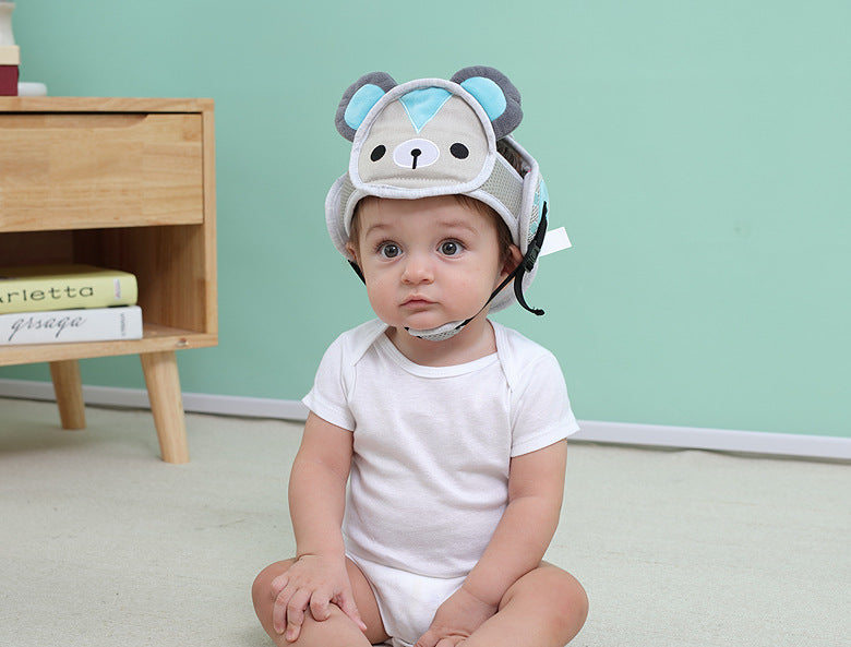A baby wearing a cute, bear-themed protective helmet and white onesie sits on the floor. A small wooden table with books is in the background against a mint-green wall.