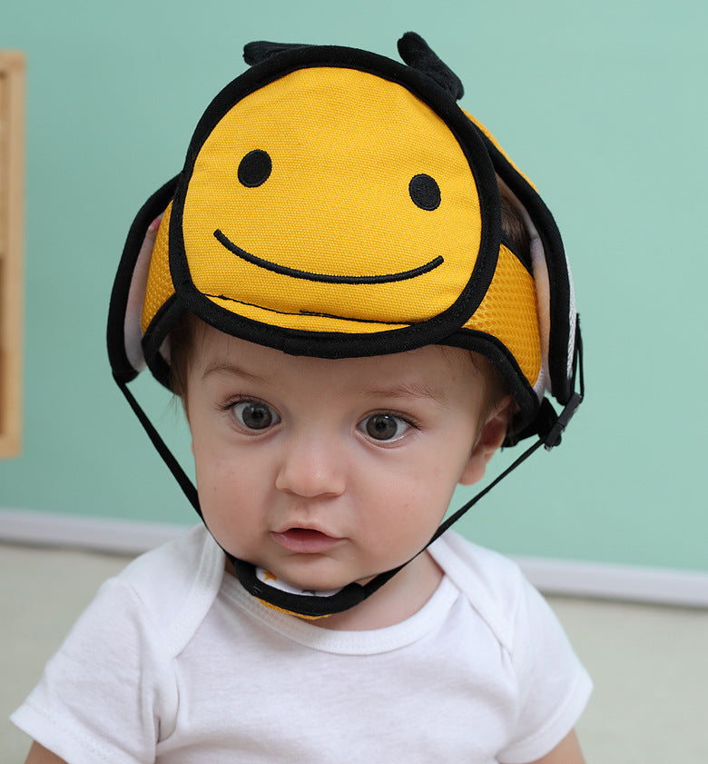 A baby wearing a yellow helmet with a smiling face design sits against a pastel green background, looking curiously forward. The helmet features black straps.