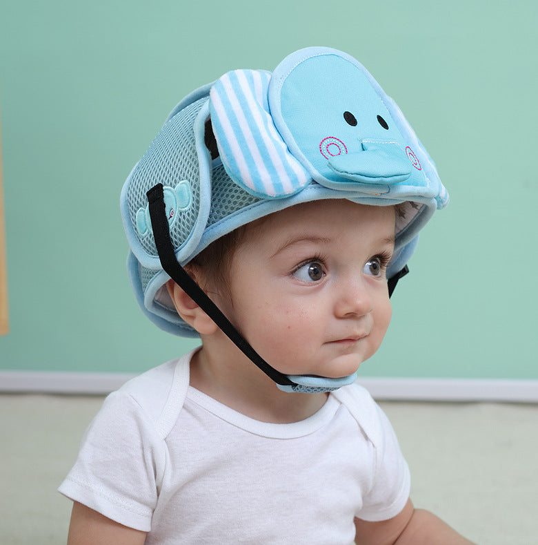 A baby in a white shirt sits against a mint green background, wearing a cute blue helmet with an elephant design featuring ears and a trunk, looking to the side.