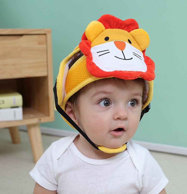 A baby wearing a cute lion-themed protective helmet with a colorful lion face design, sitting indoors with a piece of wooden furniture and books in the background.